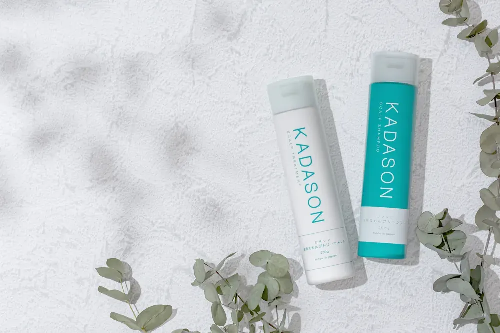 白い壁の前に置かれたKADASONのシャンプーとトリートメント、ユーカリの葉